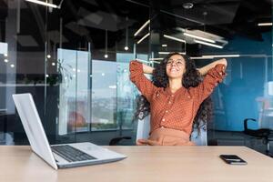 Happy and smiling successful hispanic business woman finished work satisfied with work result and achievement, hands behind head and smiling close up. photo