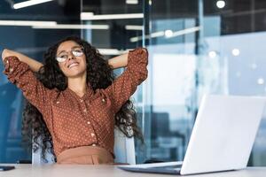 Young woman engineer developer programmer works in the office inside the IT company, satisfied with the work result, rests hispanic looks out the window, smiles hands behind her head, dreams. photo