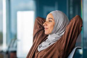 Successful completion of the project, working day. A young Arab woman in a hijab is resting at the workplace, thoughtfully and contentedly looking out the window. photo