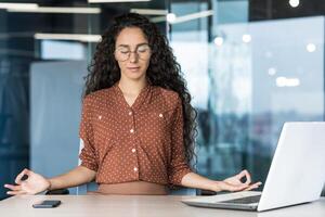 Young beautiful Hispanic web developer working inside office building, woman resting meditating at workplace with eyes closed, female worker using laptop to write code. photo