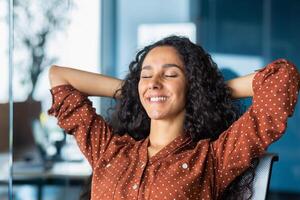 Happy and smiling successful hispanic business woman finished work satisfied with work result and achievement, hands behind head and smiling closeup. photo