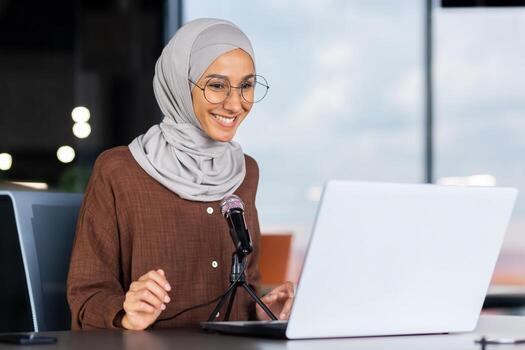 A young Muslim woman in a hijab is sitting in an office in front of a laptop and a microphone. records a podcast, a blog, talks on a call, gives an interview, goes through an online interview. photo