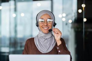 Close-up photo. Young hispanic woman in hijab sitting in office in headset and talking, consulting, advising on call from laptop. He holds the microphone in his hand, looks at the camera. photo