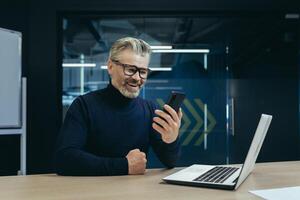 Senior smiling gray-haired male businessman sitting in the office at the desk and using a mobile phone. Reads news, messages, talks via call. photo