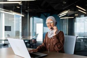 Helpline. Young hispanic woman in hijab sitting in office in headset and talking, consulting, advising on call from laptop. He holds the microphone in his hand, looks at the camera with a smile. photo