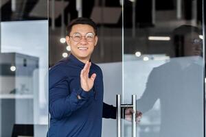 Successful asian worker smiling and looking at camera, man opens door walks into middle of meeting room, holds hand up greets colleagues. photo