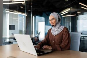 A young Muslim woman in a hijab and a headset is working in the office at a desk using a laptop. Typing on the keyboard, talking via call, consulting. photo