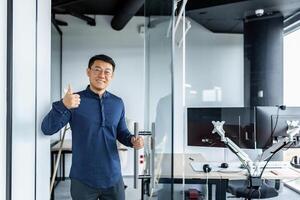 Portrait of Asian designer engineer, man smiling and looking at camera holding thumb up affirmative, businessman working with several computers inside twisted office building. photo