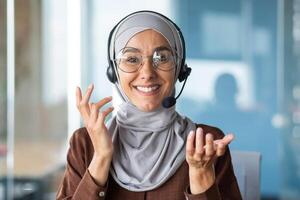 Online customer support worker consults a client remotely, a woman in a hijab with a headset for a call talks, looks at the web camera and smiles at the interlocutor, closeup photo