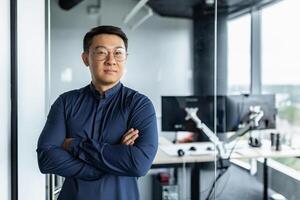 Portrait of businessman with arms crossed, asian man smiling and looking at camera, man working inside modern office building. photo