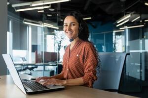 Portrait of Latin American business woman, office worker looking at camera and smiling, using headset and laptop for remote online communication, customer support tech call center worker. photo