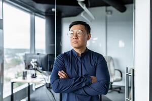 Thinking businessman with crossed arms in glasses looking out window, asian developer working inside modern office building. photo