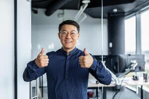 Portrait of successful asian businessman, man working in modern office looking at camera and smiling, broker trader showing thumbs up. photo