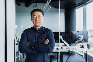 Portrait of successful Asian programmer, team leader company owner smiling and looking at camera with crossed arms, talented startup entrepreneur working inside IT office photo
