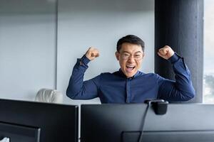 Happy Asian boss leader of programmer team, looking at computer screen and happy, celebrating victory, completed project, and submitted completed program, businessman entrepreneur holding hands up. photo