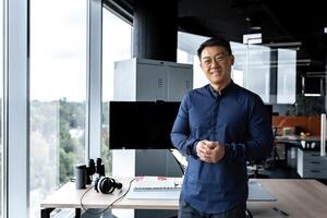 Programmer in glasses and shirt smiling and looking at camera, Asian man working inside modern computer company, working in office with several computers and monitors. photo