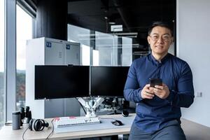 Portrait of successful businessman, man holding smartphone, asian man working inside modern office building using dual computer, worker looking at camera and smiling in glasses. photo