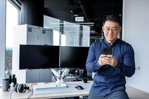 Korean worker programmer developer working inside modern office building with dual computers, man using phone, typing message, Asian man reading good news online from smartphone. photo