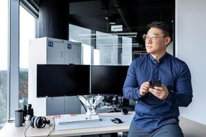 Portrait of serious Asian architect businessman, man looking out window working in modern bright office inside using dual computer, businessman holding phone in hands. photo