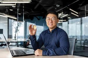 Portrait of satisfied asian broker investor, businessman looking at camera and smiling man holding crypto currency bitcoin virtual money in hands, working inside modern office building. photo