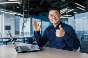 Successful asian businessman smiling and looking at camera, holding bitcoin coin and giving thumbs up, man working inside modern office building using laptop, broke stock exchange photo