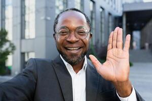Close-up photo. An African American businessman in a suit and glasses stands outside an office center and talks on a call. He is holding a phone, waving at the camera, smiling. photo