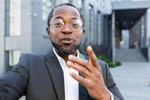 Close-up photo. African American businessman in a suit and glasses stands outside an office center and talks on a call from the phone with partners, clients. Explains, waves his hand. photo