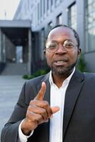 Young african american businessman, broker, real estate agent standing near office. Talks via call with partners and clients. Seriously explains, tells, shows. Shows attention index finger. photo
