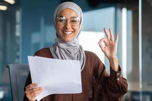 Portrait of a happy Muslim woman in a hijab holding a document, a letter, a sheet of paper. Got good news, new job, promotion. He smiles and points to the camera with his hand ok. photo