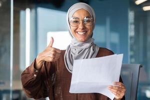 Portrait of a happy Muslim woman in a hijab, a student holding a document, a letter, a sheet of paper in her hands. Received good news, smiles and points to the camera with a super hand. photo