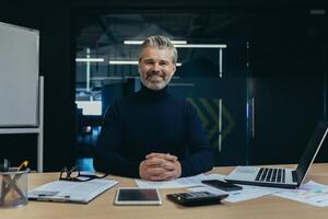 Portrait of a self-confident successful senior man businessman, accountant, manager sitting in the office at the table, smiling and looking at the camera, hands folded together. photo