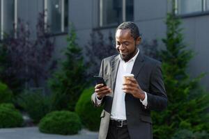 Mature businessman in business suit using phone outside office building, african american boss holding cup of hot drink, smiling typing text message and browsing websites. photo