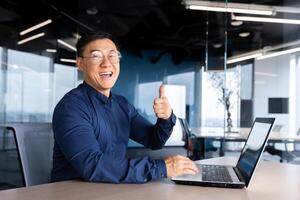 Happy and satisfied asian programmer looking at camera and smiling holding thumb up, businessman working inside office using laptop at work. photo