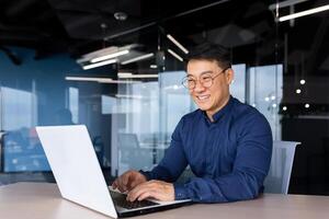 Cheerful and smiling Asian man working inside office, businessman in shirt and glasses using laptop, engineer developer programmer typing code programming software. photo