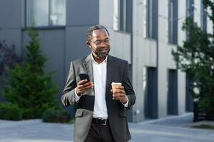 Coffee break. A successful African American businessman in a business suit is standing on the street near a skyscraper in a suit and glasses, holding a cup of coffee to go and using a mobile phone. photo