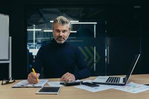 Portrait of a senior man businessman accountant, financial director working in the office at a table behind a laptop and with documents. Confidently looks into the camera. photo