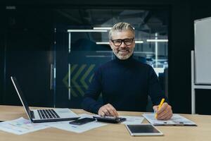 Portrait of an older gray-haired man, an accountant, an auditor sitting in the office at a laptop and a calculator. He works with documents, checks accounts. Smiling at the camera. photo