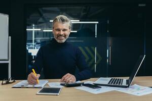 Portrait of a confident senior man businessman, accountant, manager sitting in the office at the table, smiling and looking at the camera. Work by laptop and with documents. photo