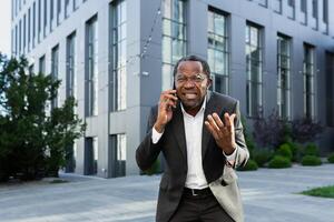 Angry african american boss arguing and shouting while talking on the phone, senior man in business suit outside office building. photo