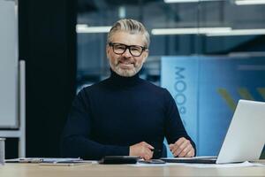 Senior portrait of a gray-haired businessman, founder, executive director of the company, who works in the office with a laptop and documents. Smiling and confidently looking at the camera. photo