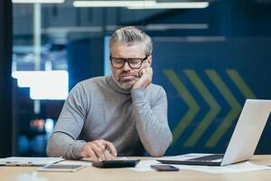Upset and disappointed senior and mature financier, man working inside office building, calculating financial report with calculator, gray haired businessman investor in glasses thinking paperwork photo