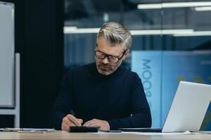 An older male businessman sits in the office at a table with a laptop, writes documents. Working on a project, deadline, overtime, at night time. photo