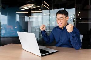 A happy young Asian student man sits in the office at the table, looks at the laptop and rejoices. Passed the exam, received a letter, accepted a job, entered the university. Screams with joy. photo