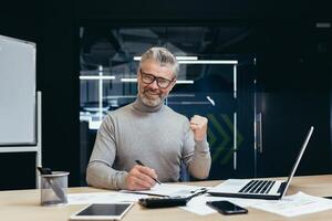 Portrait of successful mature financier, senior gray-haired man looking at camera and smiling, businessman in glasses paper work on reports and deals, using calculator and notebook in office. photo