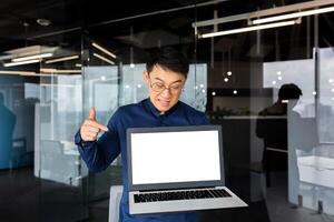 Worried and upset young Asian man, programmer, businessman, freelancer standing in the office and holding a broken laptop. Pointing an angry finger at an illuminated blank screen. photo