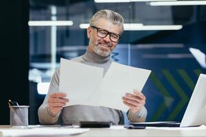 Senior smiling gray-haired businessman working in office at desk with laptop and documents. Holds in his hands, looks at sheets of paper, agreements, bills. photo