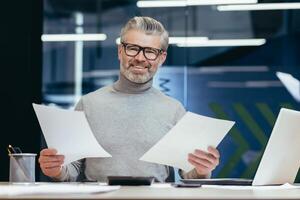 Successful businessman with documents smiling and looking at camera, portrait of mature gray-haired investor financier inside modern office, man behind paper work with reports accounts and contracts photo