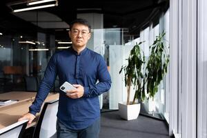 Portrait of serious thinking asian man inside office, man in glasses and shirt looking at camera, mature programmer software developer at workplace holding phone near window. photo