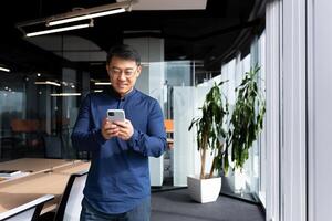A young man, an Asian businessman, a student standing by the window in the office, uses a mobile phone, types messages, reads, checks mail. He looks at the camera, smiles. photo