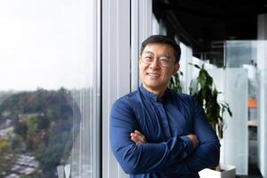 Portrait of successful asian boss inside office, businessman with crossed arms smiling and looking at camera, man in shirt and glasses happy with result of achievement at work. photo